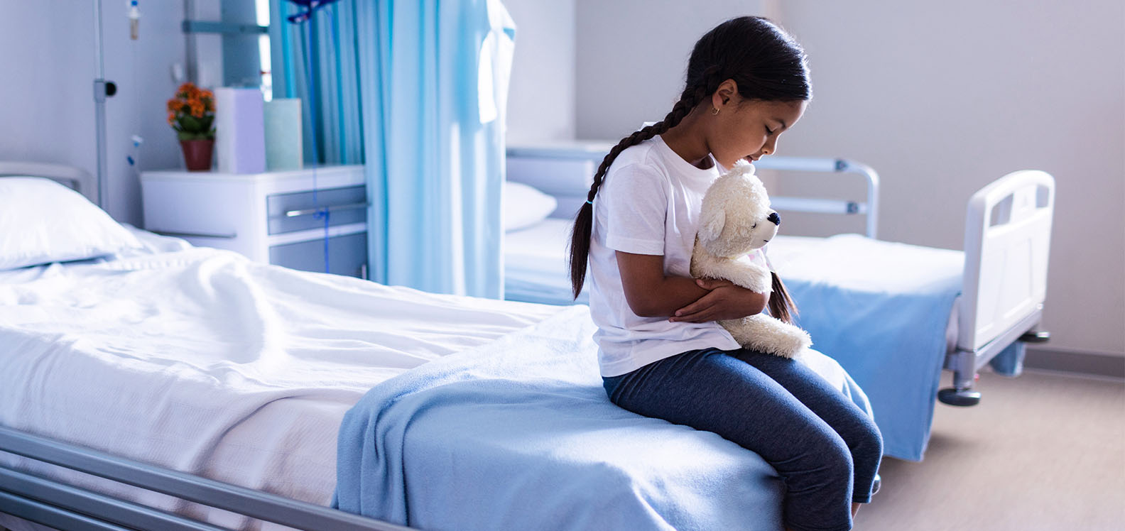 Female child holding a soft toy sitting on a hospital bed in a ward.