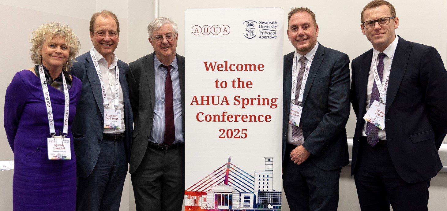 Five smartly dressed people - four men and one woman - standing alongside a conference pop-up banner.