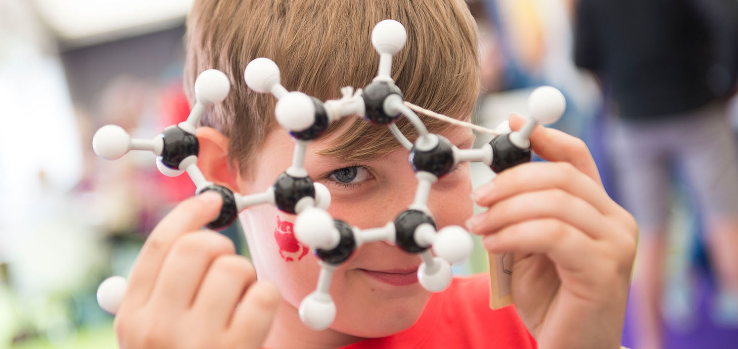 A child holds a molecular model in front of their face, looking through the structure, with blurred people and objects in the background.