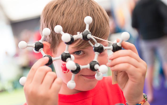 A child holds a molecular model in front of their face, looking through the structure, with blurred people and objects in the background.