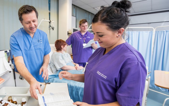 Nurses on a ward by an open medicines trolley with patient in hospital bed in background