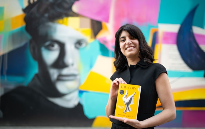 Yasmin Zaher stands holding a copy of her novel,The Coin, in front of a colourful mural featuring the face of Dylan Thomas. 