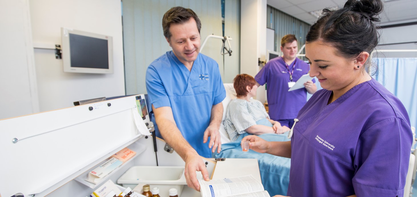 Nurses on a ward by an open medicines trolley with patient in hospital bed in background