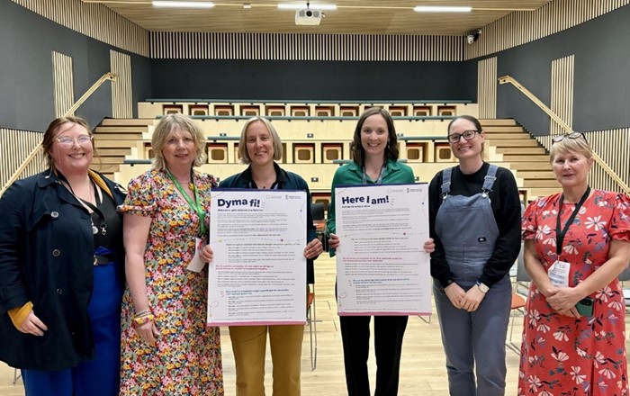 Six women standing side by side in front of an auditorium, two holding placards
