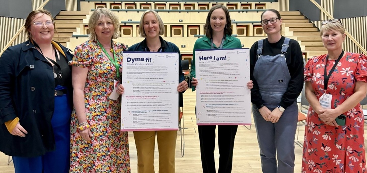 Six women standing side by side in front of an auditorium, two holding placards