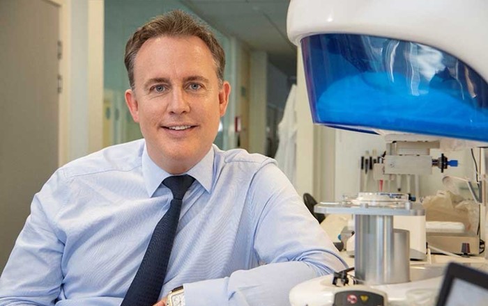 White formally dressed man, leaning against lab equipment, looking at camera.