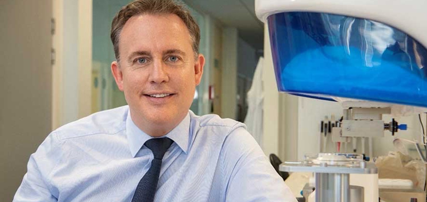 White formally dressed man, leaning against lab equipment, looking at camera.