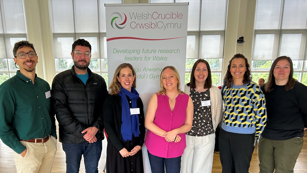 A group photo of the seven Swansea University researchers standing together and smiling in front of a branded pull-up banner displaying the Welsh Crucible logo.