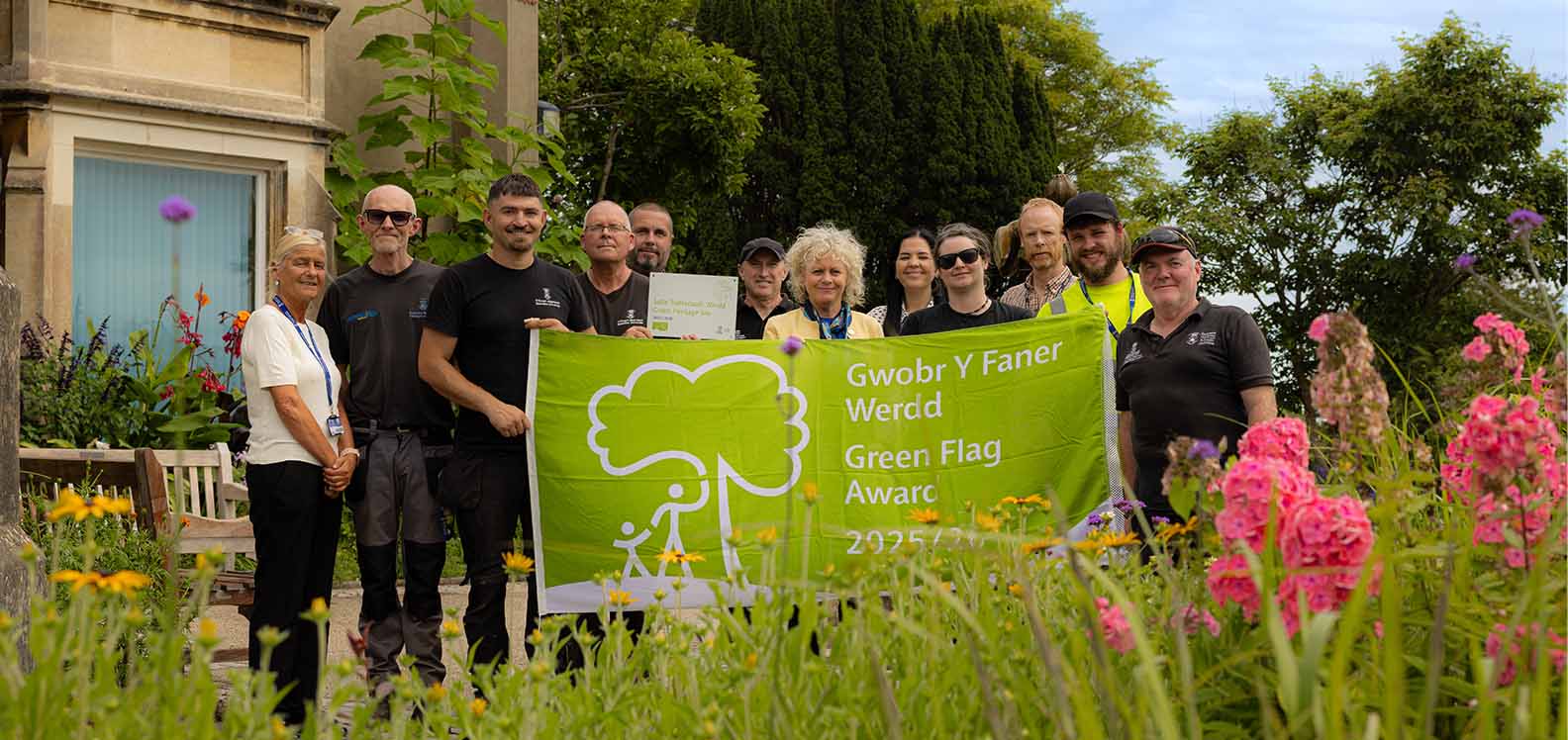 Group of people standing outside a building holding a green flag, surrounded by flowers.