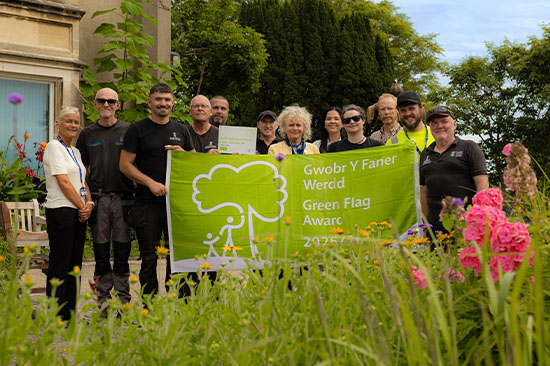 Group of people standing outside a building holding a green flag, surrounded by flowers.