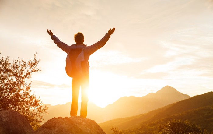 An example of someone thriving: A hiker stands on a mountain cliff with a backpack. The sun rises behind them. Credit: lenina11only | Shutterstock.