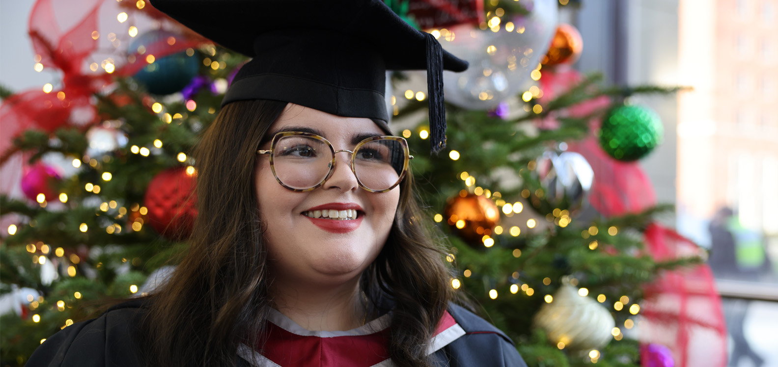 Young woman in graduation robes standing in front of a decorated Christmas tree