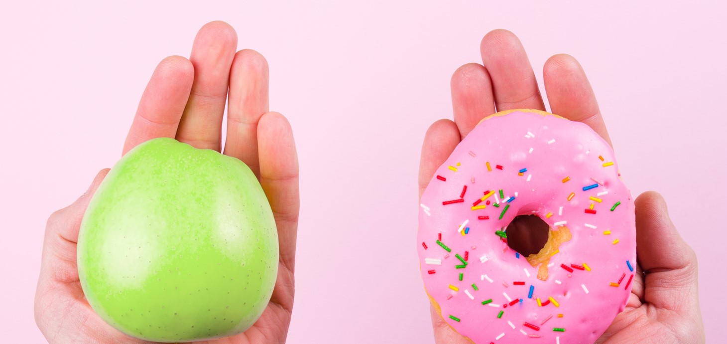 Against a pale pink background, one hand is holding a green apple and another hand is holding a pink glazed donut with multicoloured sprinkles. 