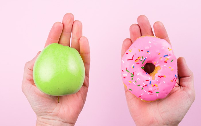 Against a pale pink background, one hand is holding a green apple and another hand is holding a pink glazed donut with multicoloured sprinkles. 