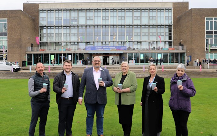 Six people standing on grass in front of a building holding reuseable coffee cups