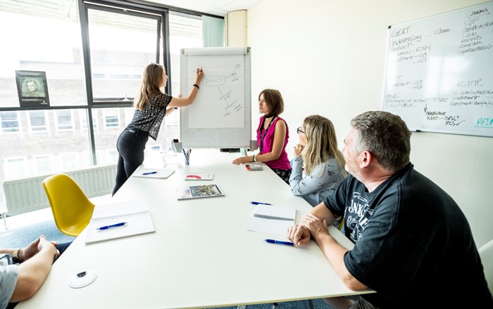 Woman writing on white board in front of three other people in a meeting room.