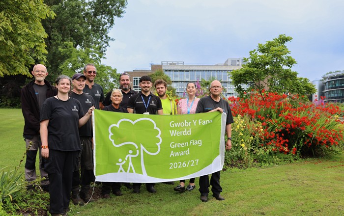 Group of people holding up large green flag in front of a flower border. 