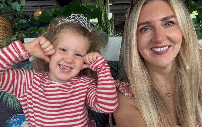 Smiling small girl wearing toy tiara sitting indoors alongside smiling adult woman.