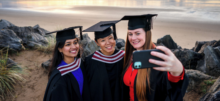 Three females dressed in graduation robes pose for a selfie on the beach 