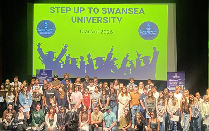A large group of young people gathered on stage in front of a large green screen that reads 'Step Up to Swansea University' 