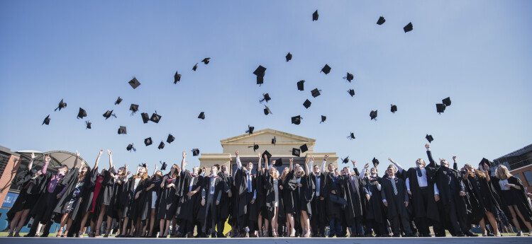 A large group of students in graduation robes throw their mortarboards in the air while standing in front of Swansea University’s Great Hall.