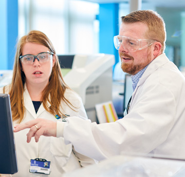 Man and woman in lab coats and goggles discussing something on a screen