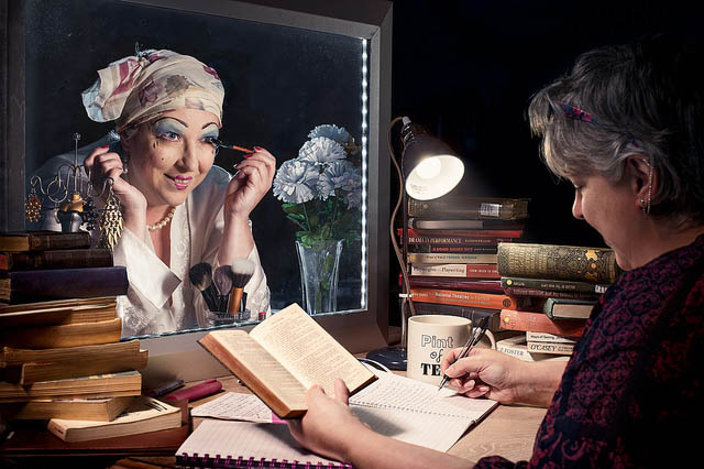 A woman reading books in front of a mirror, her reflection is putting on make-up