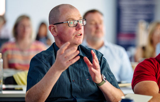 A man in a dark shirt speaking to from his seat