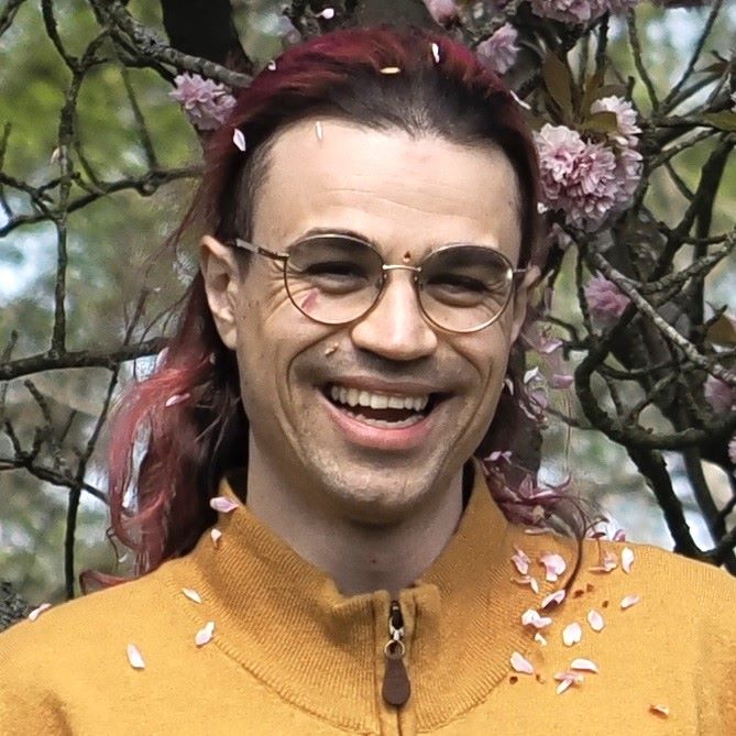  person (troy) with pink hair and glasses smiles as pink petals fall from the cherry tree above across their face and onto their mustard-coloured cardigan