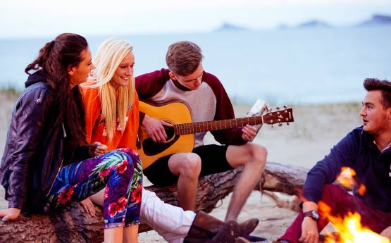 Friends gathered around a campfire by the beach with a boy playing guitar
