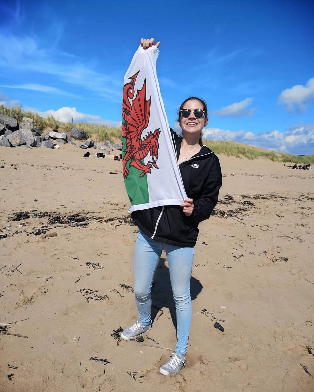 Student wearing sunglasses holding Welsh flag on Swansea beach