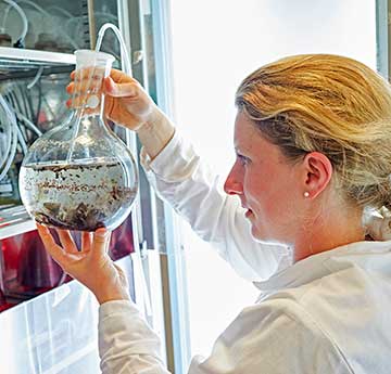 An image of a female in a white lab coat in a laboratory holding a large glass cylinder with a liquid and sediment inside. 
