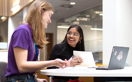 An image of two students talking at a table in a library