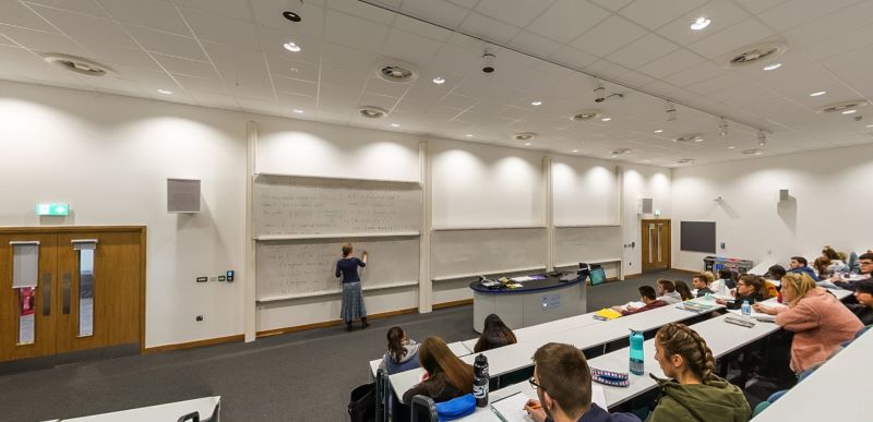 Lecture theatre with tiered seating, lecturer writing on boards, and students ta