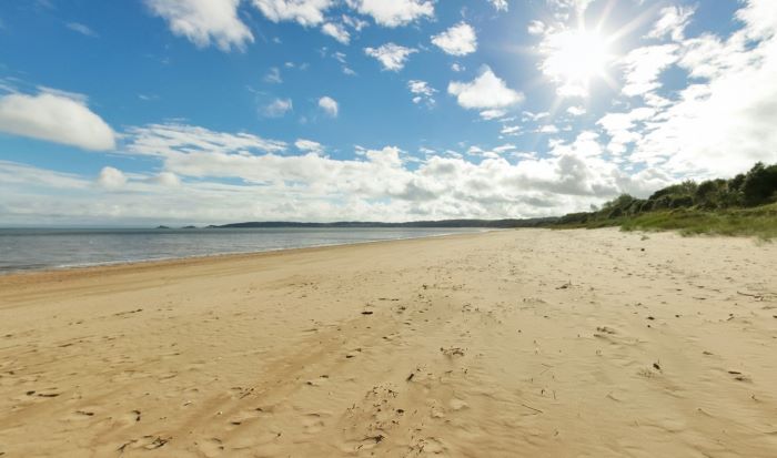 Wide sandy beach with calm sea, blue sky, scattered clouds, dunes